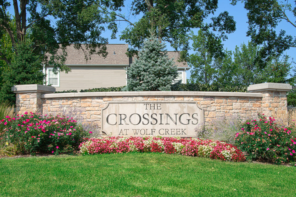 Stone entrance sign for The Crossings at Wolf Creek surrounded by flowers, trees, and lawn
